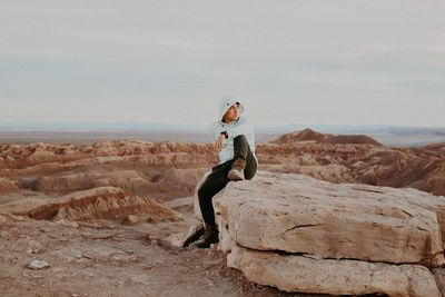 Full length of young man sitting on rock formation against sky during sunset