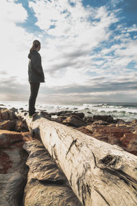 Side view of woman standing on log by sea against cloudy sky