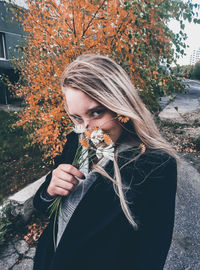 Portrait of young woman holding flowers while standing against autumn tree