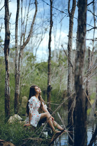 Thoughtful young woman sitting at lakeshore surrounded with bare trees