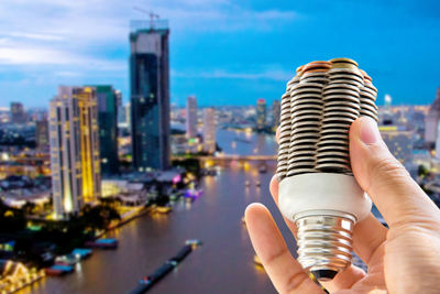 Cropped image of hand holding light bulb made of coins against cityscape