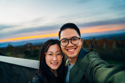Portrait of smiling couple taking selfie against sky during sunset