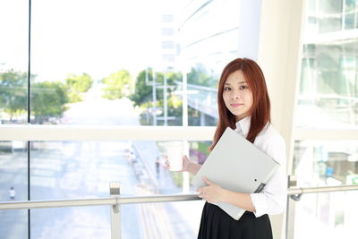 Portrait of young woman holding file while standing in office corridor