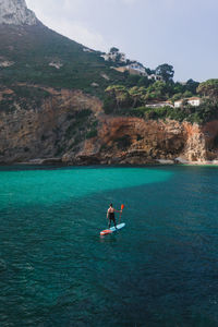 Back view of full body male surfer balancing on paddleboard with oar in hands while practicing sup surfing on turquoise seawater in sunny day