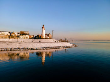 Lighthouse by sea against buildings against clear blue sky