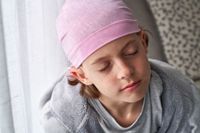 Serious cute child in pink bandana with closed eyes fighting cancer at home sitting in a couch