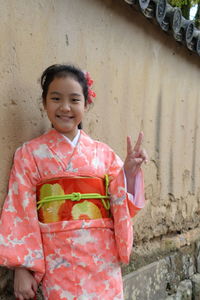 Portrait of smiling girl wearing kimono gesturing peace sign while standing against wall
