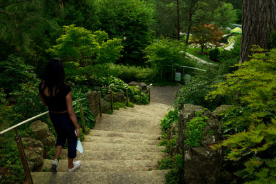 Rear view of woman walking with mobile phone and protective face mask on steps amidst trees