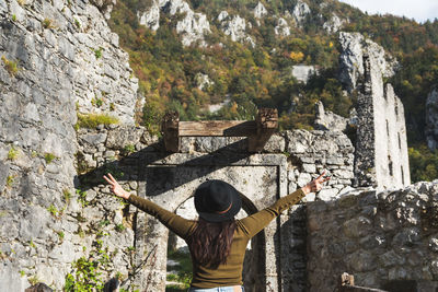 Rear view of woman in autumn clothes standing in old castle ruin, arms outstretched.