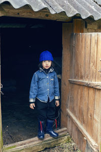 Village boy in boots stands at the door of the barn in warm clothes