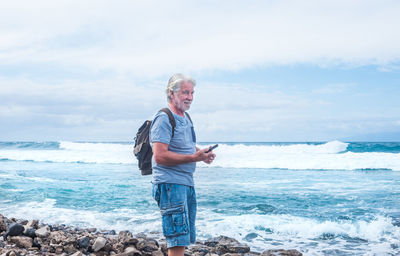 Side view of backpack senior man with phone standing at beach against sky