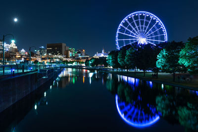 Illuminated ferris wheel by river against sky in city at night