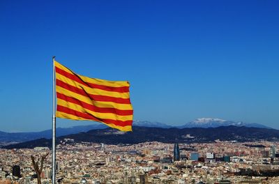 Catalan flag against buildings of barcelona cityscape and background of mountains against blue sky