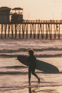 Woman carrying surfboard at sunset by pier