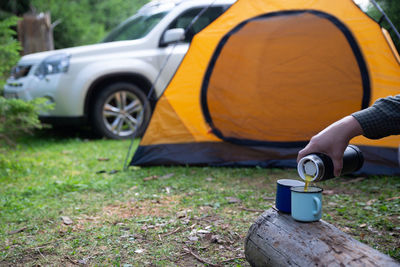 Man hand pour tea in metal cup. yellow tent with suv on background. union with nature