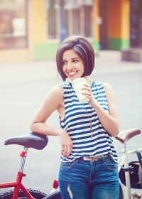 Smiling young woman drinking coffee while standing by bicycle