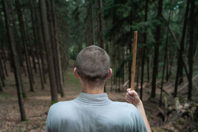 Back view of unrecognizable man with stick standing on rock near trees while practicing kung fu in coniferous forest