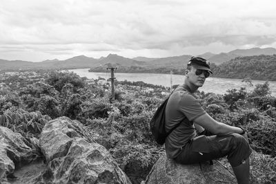 Side view of man relaxing on rock against river and mountains