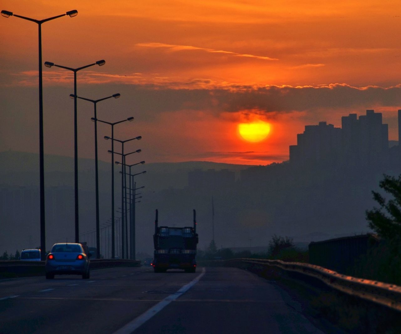 sunset, orange color, transportation, car, sky, road, building exterior, land vehicle, silhouette, mode of transport, architecture, built structure, cloud - sky, city, street, sun, street light, dramatic sky, the way forward, nature