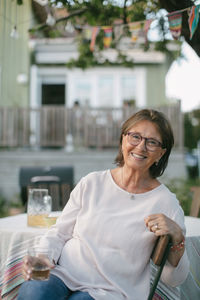 Portrait of smiling senior woman holding drink while sitting on chair at table in backyard