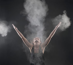Close-up of woman with arms raised standing amidst talcum powder against black background