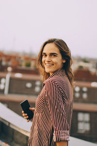 Portrait of happy young woman holding mobile phone standing on terrace in city