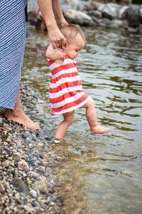Close up view on baby in striped dress making steps in water being held by motherhands