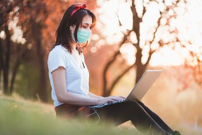Side view of woman wearing flu mask using laptop at park