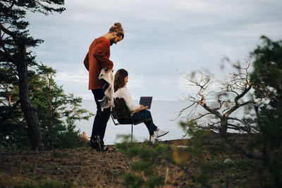 Man holding blanket while woman using laptop in forest against sky