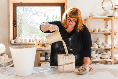 Adult craftswoman in black apron and casual clothes standing near table and pouring clay into a mold near bucket and window near shelves with earthenware in workshop in daytime