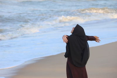 Young woman wearing hijab with arms outstretched standing on shore at beach