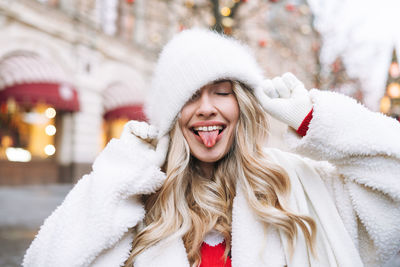 Young happy woman with curly hair in white fur hat having fun at christmas fair in winter street 
