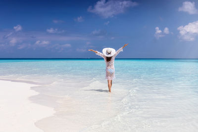 Rear view of woman with arms raised walking on shore at beach against sky during summer