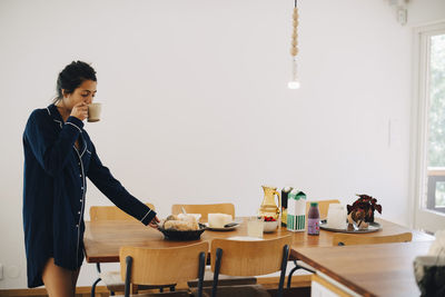 Woman drinking coffee while looking at breakfast on table by wall