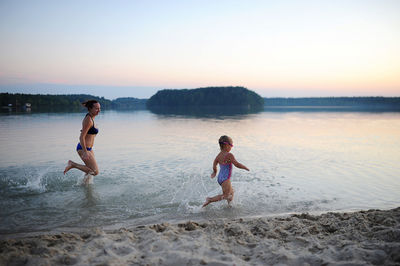 Side view of mother and daughter running at beach against sky during sunset