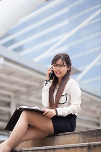 Portrait of businesswoman talking on mobile phone while sitting against office building