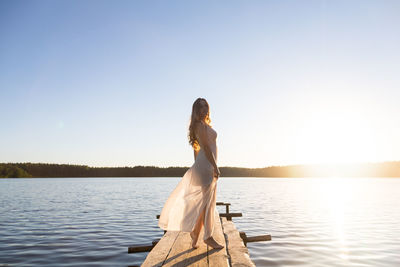 Beautiful russian girl dressed in a white dress, walking along a wooden pier on the bank of a lake