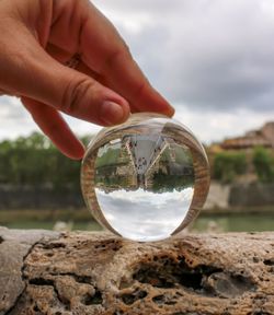 Reflection of hand holding crystal ball on rock against sky