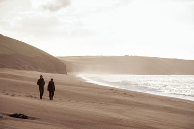 Rear view of people walking at beach against sky during sunset