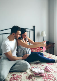 Man with pregnant women looking baby clothing while sitting on bed at home