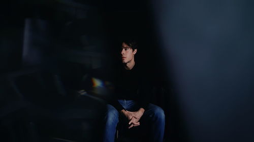 Thoughtful young man looking away while sitting on chair in darkroom