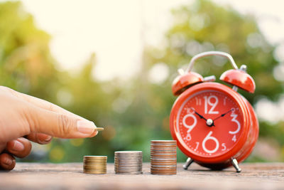 Cropped image of hand stacking coins by orange alarm clock on table outdoors