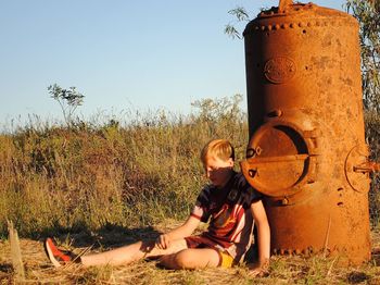 Teenage boy sitting against rusty stove on field against clear sky