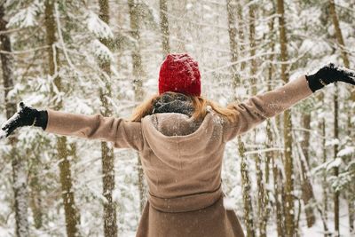 Rear view of woman with arms outstretched standing in forest during winter