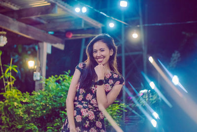 Portrait of smiling young woman standing against illuminated lights at night