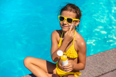 Portrait of cute girl applying suntan lotion sitting by swimming pool