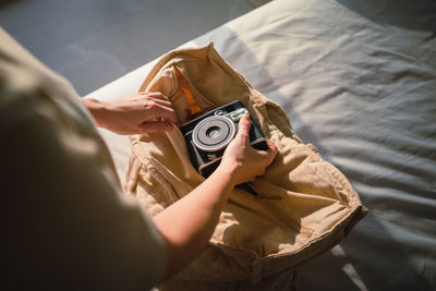 From above of crop anonymous female traveler putting vintage photo camera into cotton bag standing near bed on sunny day