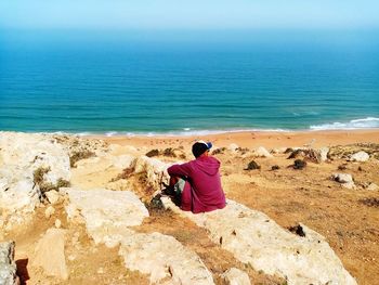 Rear view of woman sitting on rock at beach against sky