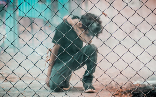 Full length of man kneeling on footpath seen through chainlink fence