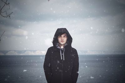 Portrait of handsome young man wearing warm clothing while standing against sea during winter